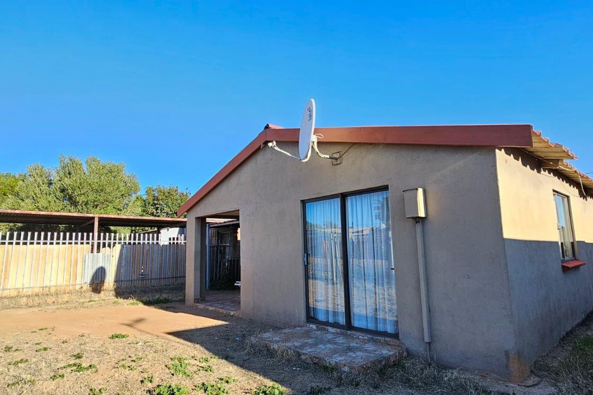 Rear view of the property featuring a simple facade, sliding glass doors, and an attached carport structure within a large, unlandscaped yard.