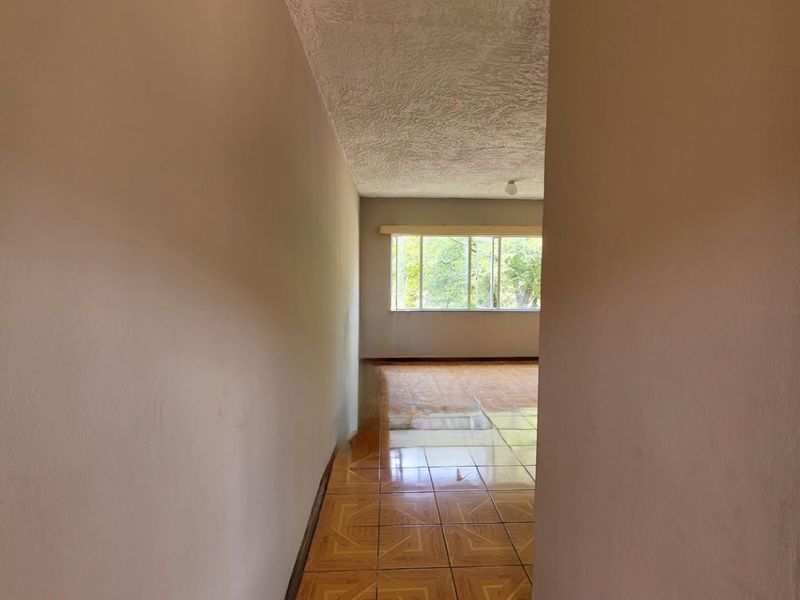 A vacant hallway with tiled flooring and a textured ceiling, leading into a naturally lit living area with a large window.