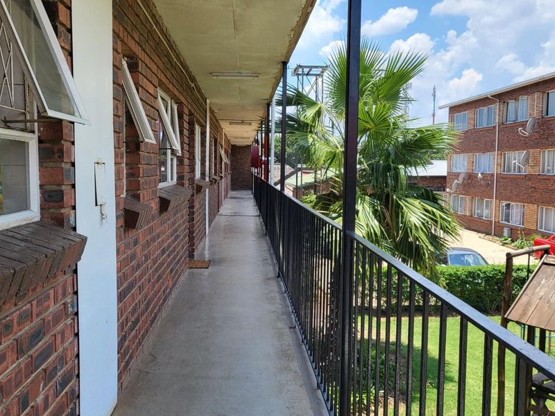 An exterior apartment balcony featuring a brick facade, metal safety railing, and concrete flooring overlooking the complex grounds.