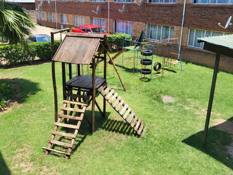 A communal garden area featuring a lawn and children's play equipment, situated within an apartment complex with brick exterior walls.