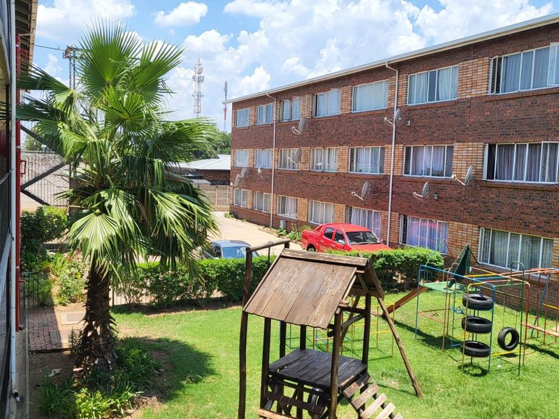 View of the apartment complex rear exterior, featuring a shared garden area, brick facade, and balcony walkways.