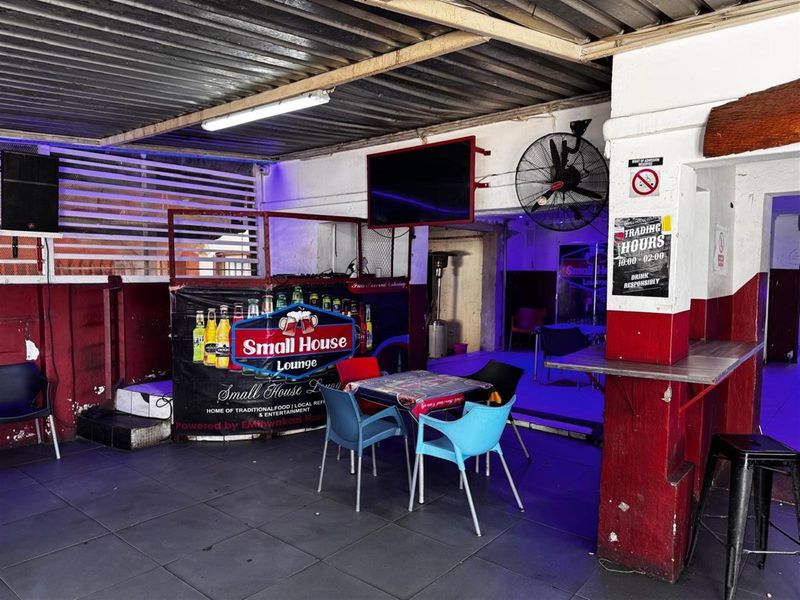 Covered outdoor patio area featuring tiled flooring, industrial wall-mounted fan, and corrugated metal roofing, currently utilized as a commercial lounge space.