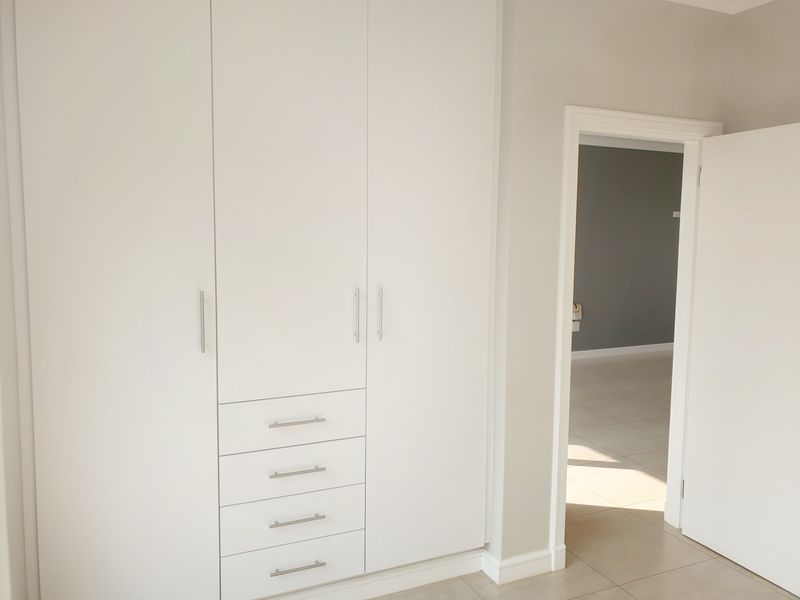 A bright bedroom featuring modern white built-in cupboards with integrated drawers and neutral tiled flooring, leading into an adjacent living space.