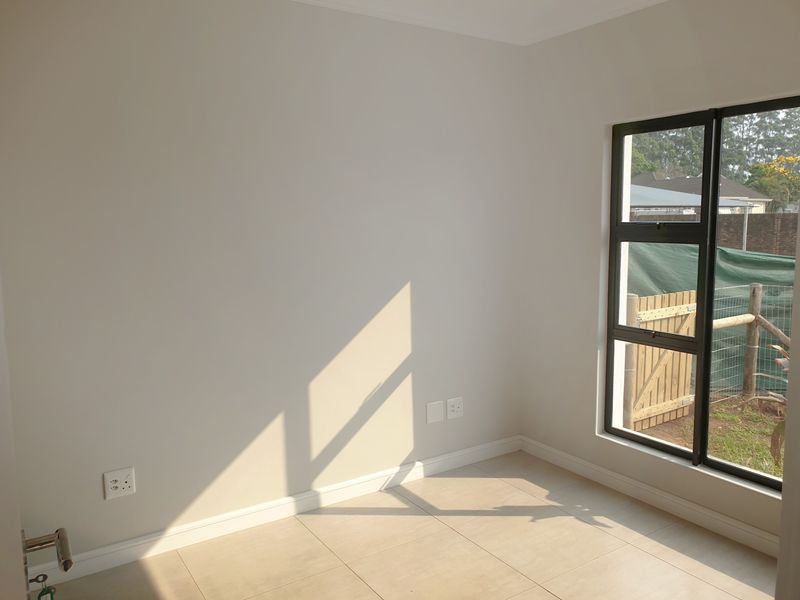 A bright, empty bedroom featuring neutral wall tones, tiled flooring, and a large aluminium-framed window overlooking the garden area.
