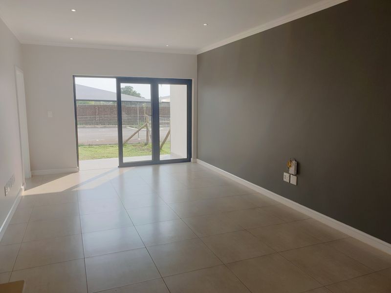 A bright, modern, and empty living room featuring neutral tiled flooring, a dark accent wall, and a large sliding glass door leading to the exterior.