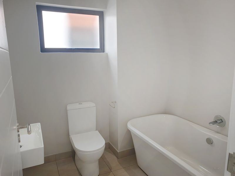 A clean, modern bathroom featuring a white freestanding bathtub, wall-mounted basin, and toilet with neutral floor tiling and a frosted glass window.