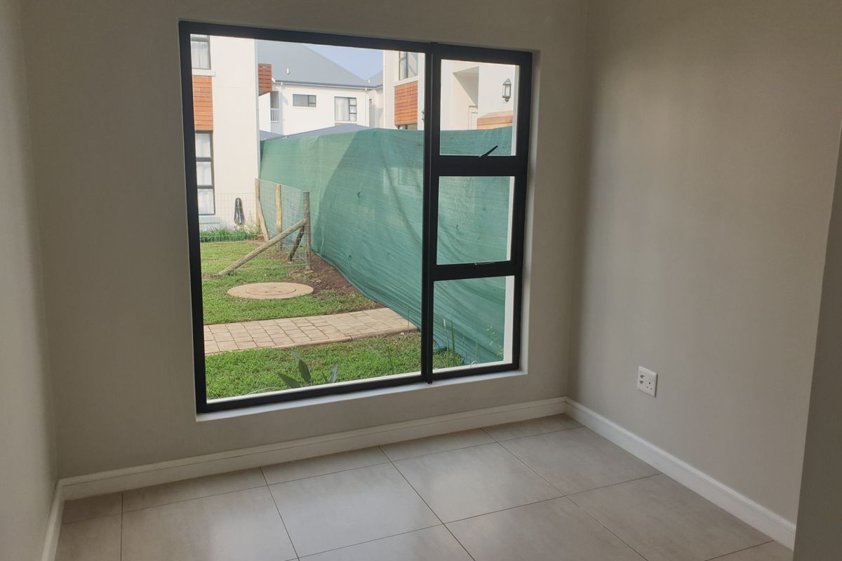 A bright, empty bedroom featuring modern tiled flooring, neutral walls, and a large aluminium-framed window overlooking the rear garden area.