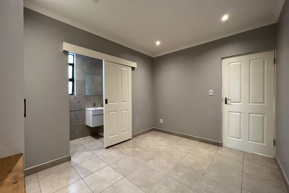 A clean, modern bedroom featuring neutral grey walls, tiled flooring, recessed lighting, and an en-suite bathroom accessed via a sliding barn-style door.