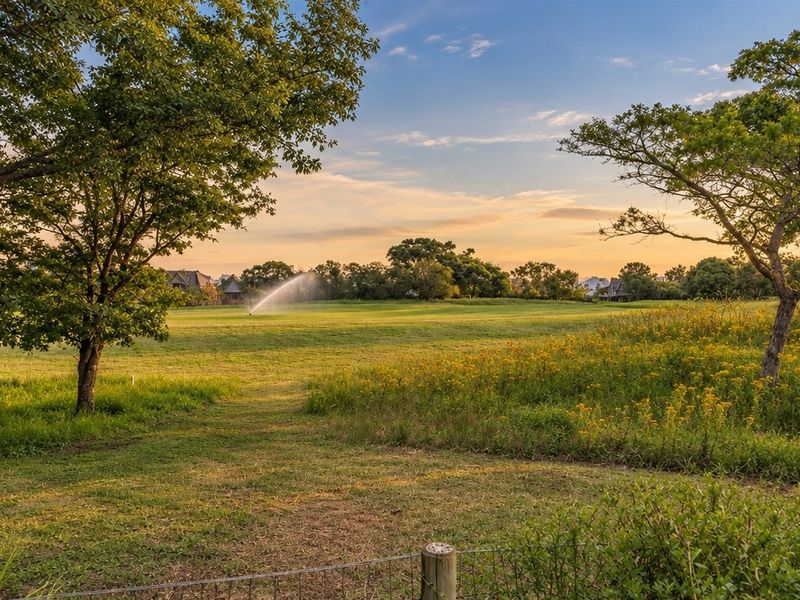 A scenic view of a well-maintained open green space with mature trees and an active irrigation system at sunset.