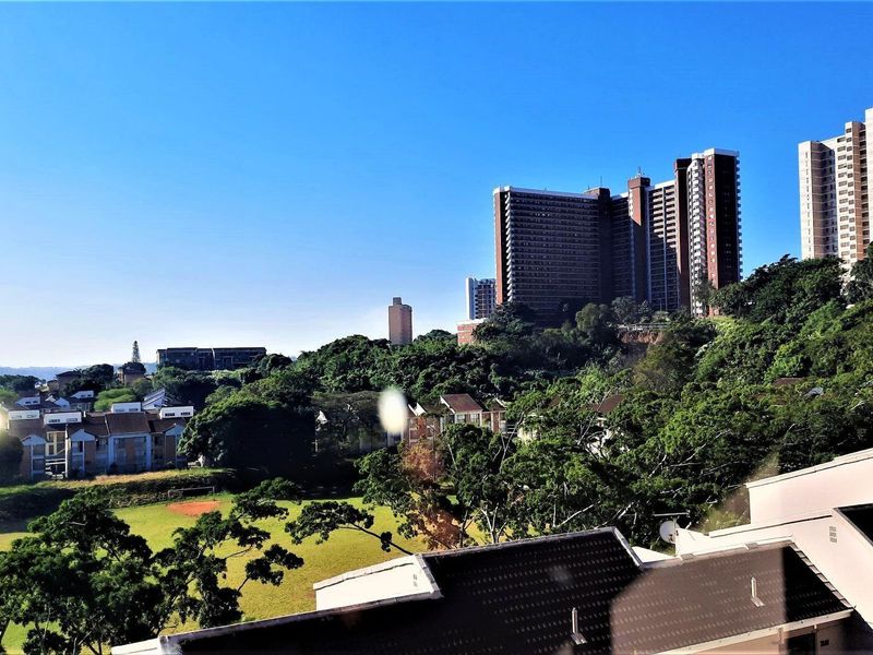 Elevated panoramic view overlooking the surrounding residential neighbourhood, lush greenery, and distant high-rise buildings under a clear blue sky.