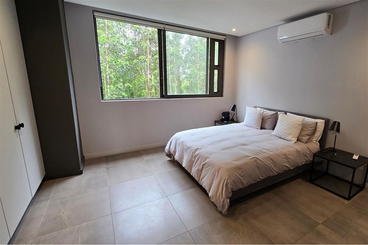A modern bedroom featuring large windows with a green outlook, tiled flooring, built-in cabinetry, and a wall-mounted air conditioning unit.