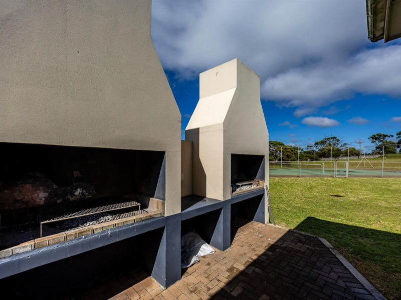 Outdoor entertainment area featuring dual built-in brick braai fireplaces on a paved patio overlooking a grassed yard and tennis court.