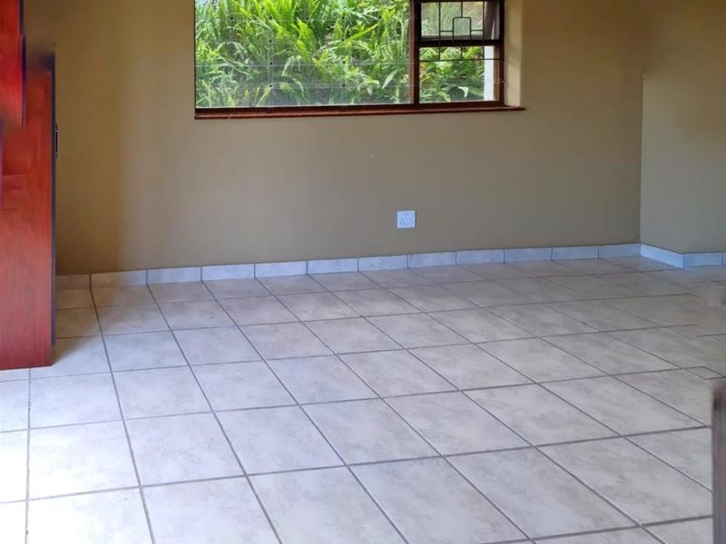 An empty living room featuring neutral tiled flooring, a large window with garden views, and simple wall finishes.