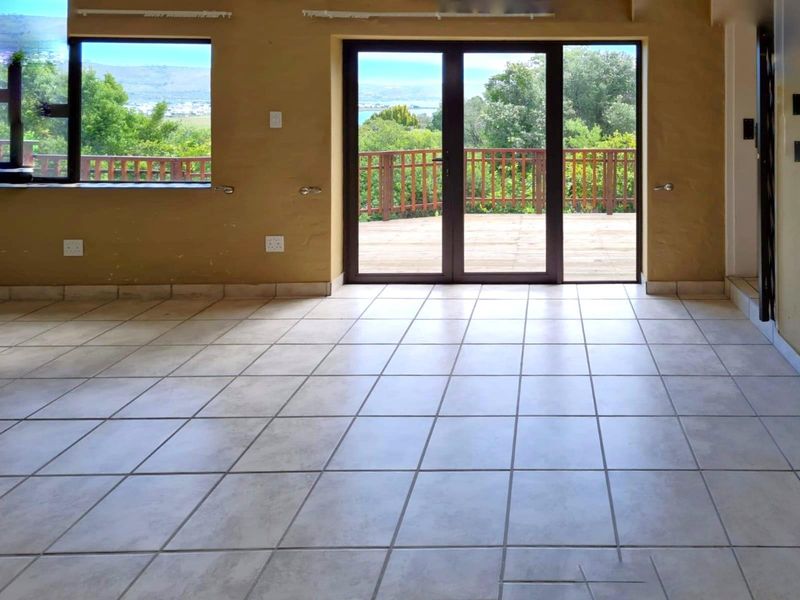 An empty living room featuring tiled flooring, large windows, and double doors leading to an outdoor deck with scenic views.