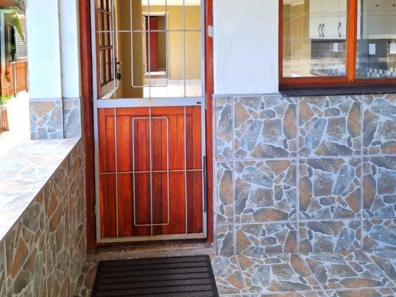 A covered patio area featuring stone-look tiled walls and flooring, a wooden security door, and a view into the interior living space.