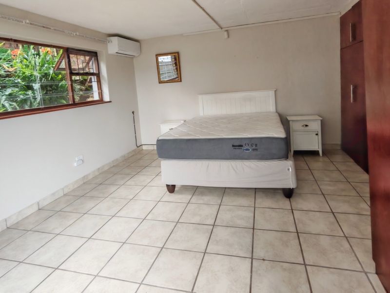 Spacious bedroom featuring tiled flooring, built-in cupboards, and an air conditioning unit, with large windows providing natural light and garden views.