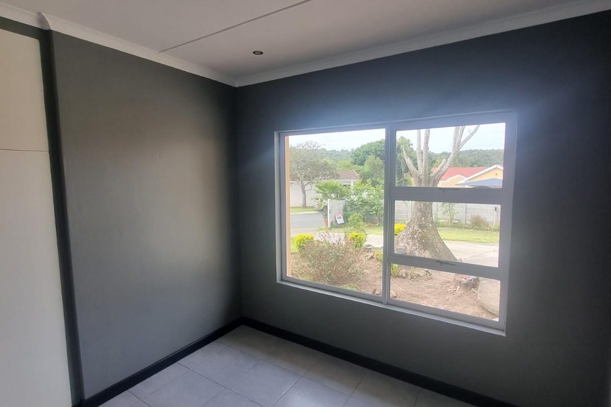 A bright, empty bedroom featuring neutral grey walls, tiled flooring, and a large window providing natural light and views of the front garden.