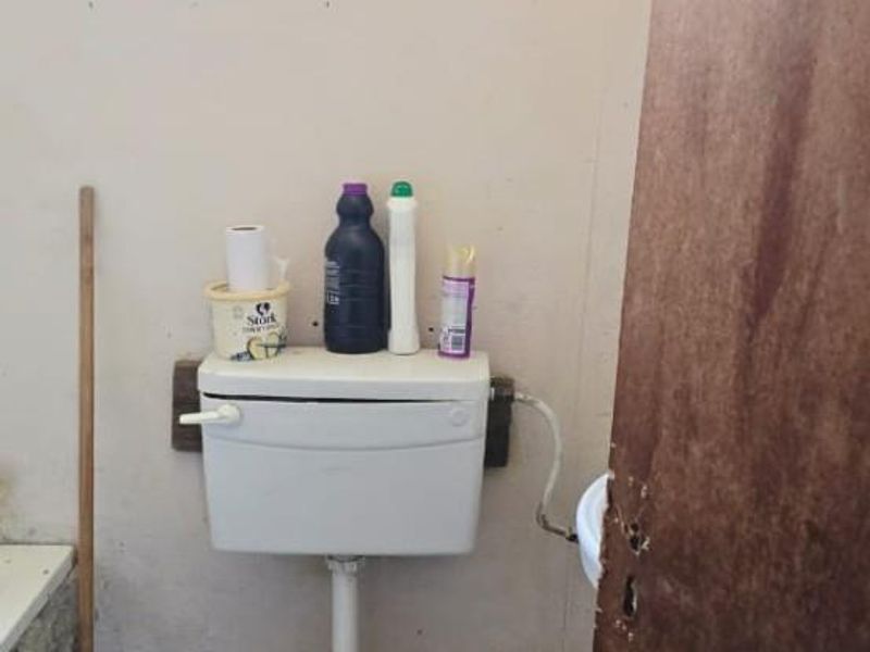 A small bathroom featuring a standard toilet, tiled flooring, and a window with security bars, requiring cosmetic updates and maintenance.