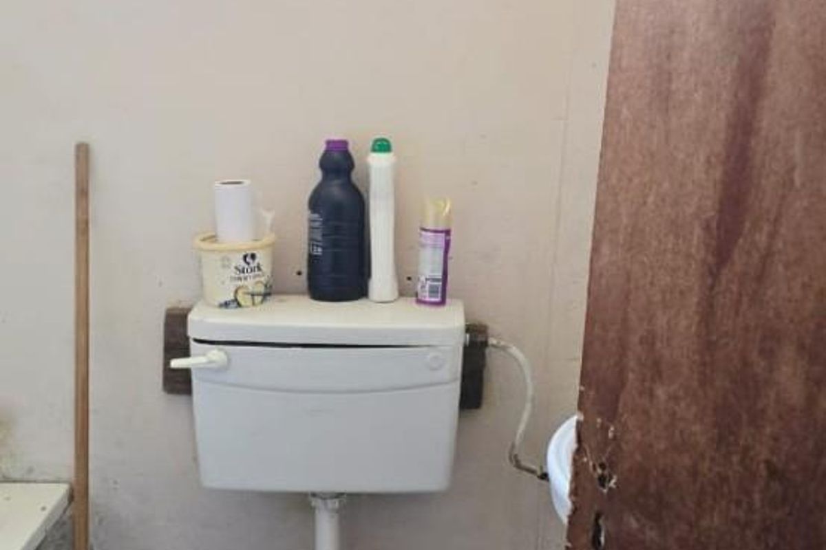 A small bathroom featuring a standard toilet, tiled flooring, and a window with security bars, requiring cosmetic updates and maintenance.