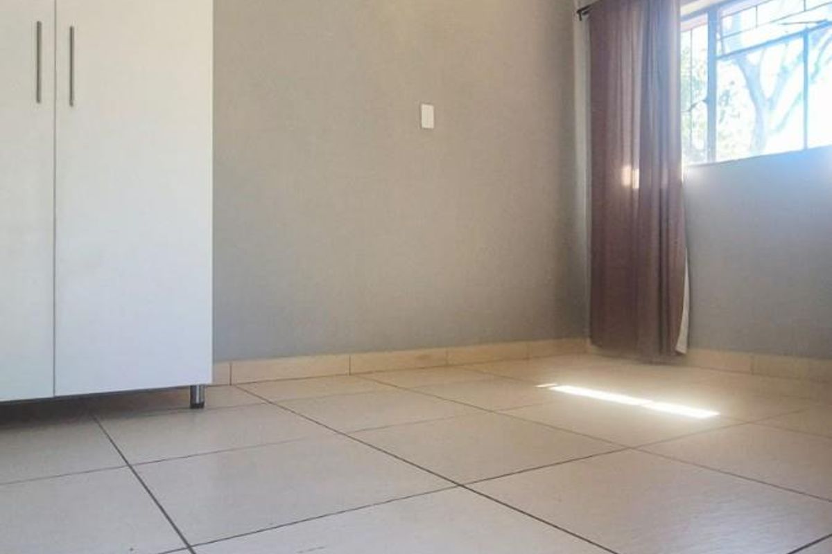 A bright, empty bedroom featuring neutral tiled flooring, a built-in wardrobe, and a large window allowing for natural light.