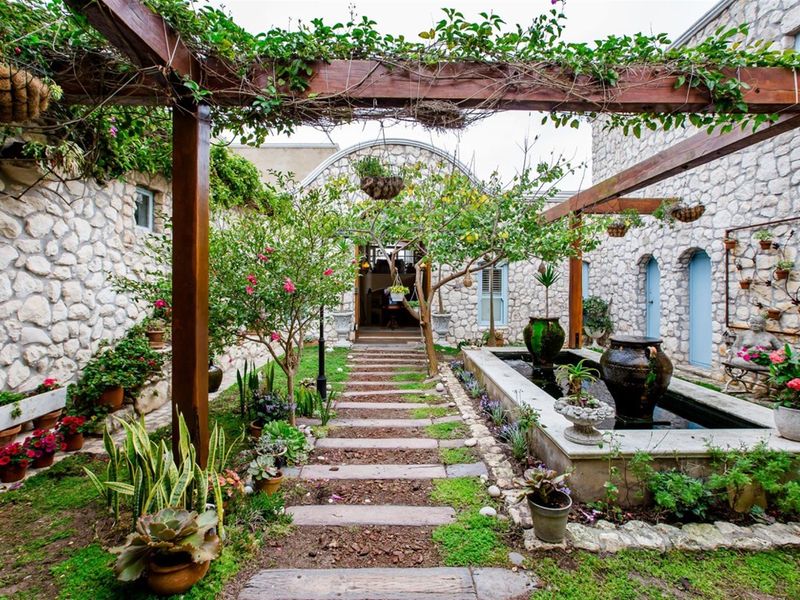 A beautifully landscaped courtyard garden featuring stone masonry walls, a timber pergola, a central stone water feature, and a stone-paved pathway.