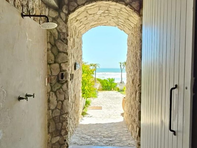 An outdoor shower area featuring a rustic stone archway, concrete flooring, and direct access to a scenic coastal garden path.