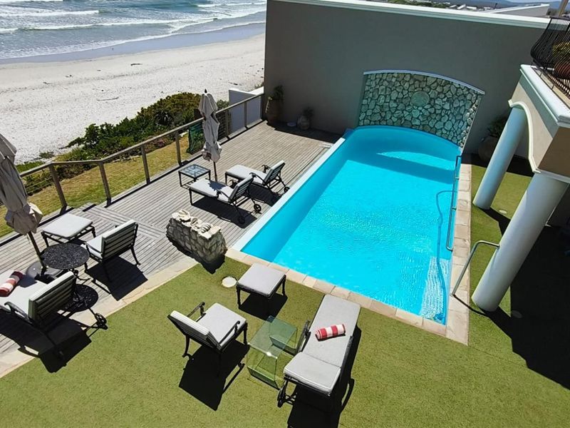 An elevated view of a private swimming pool area featuring timber decking, artificial grass, and a stone water feature overlooking the beach.
