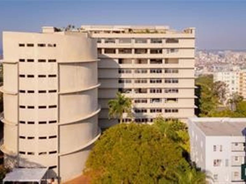 An elevated exterior view of a multi-story apartment building featuring a distinctive cylindrical architectural design and surrounding mature trees.