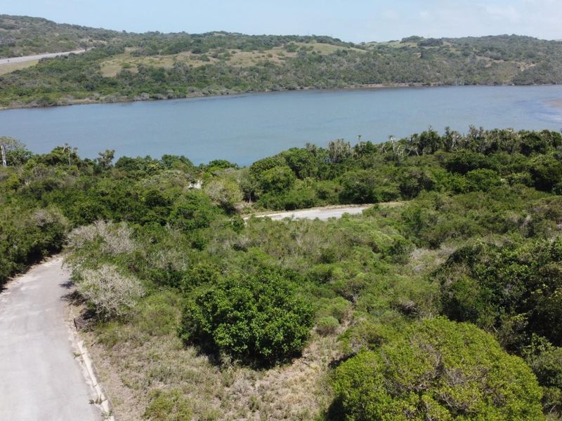 An aerial view of a vacant residential plot featuring natural vegetation, access road, and scenic views of the nearby lagoon and surrounding landscape.