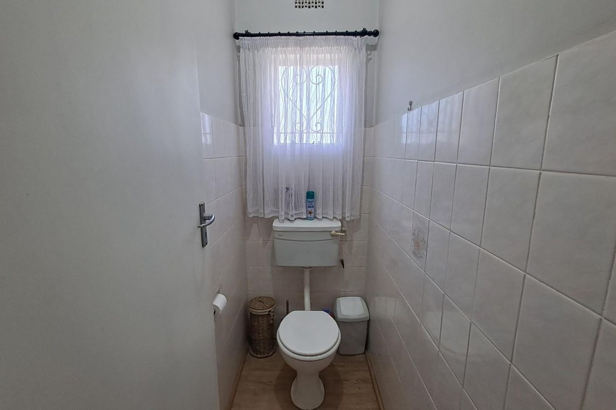 A compact, functional bathroom featuring white wall tiles, a standard toilet, and a window with security bars.