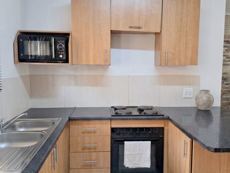 A functional kitchen featuring light wood-finish cabinetry, dark laminate countertops, a built-in oven with electric hob, and a double bowl stainless steel sink.