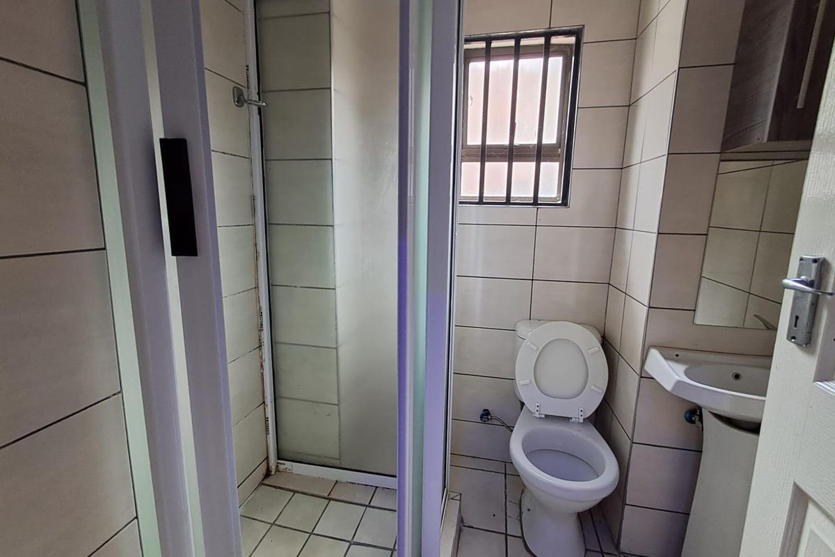 A functional bathroom featuring a shower enclosure, pedestal basin, and toilet, finished with neutral wall and floor tiling and a security-barred window.