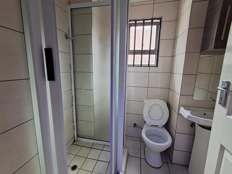 A functional bathroom featuring a shower enclosure, pedestal basin, and toilet, finished with neutral wall and floor tiling and a security-barred window.