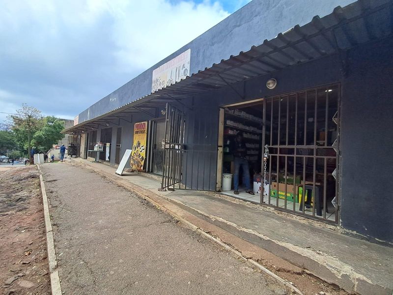 Front exterior view of a commercial strip building featuring multiple storefronts with metal security gates and a continuous corrugated metal awning.