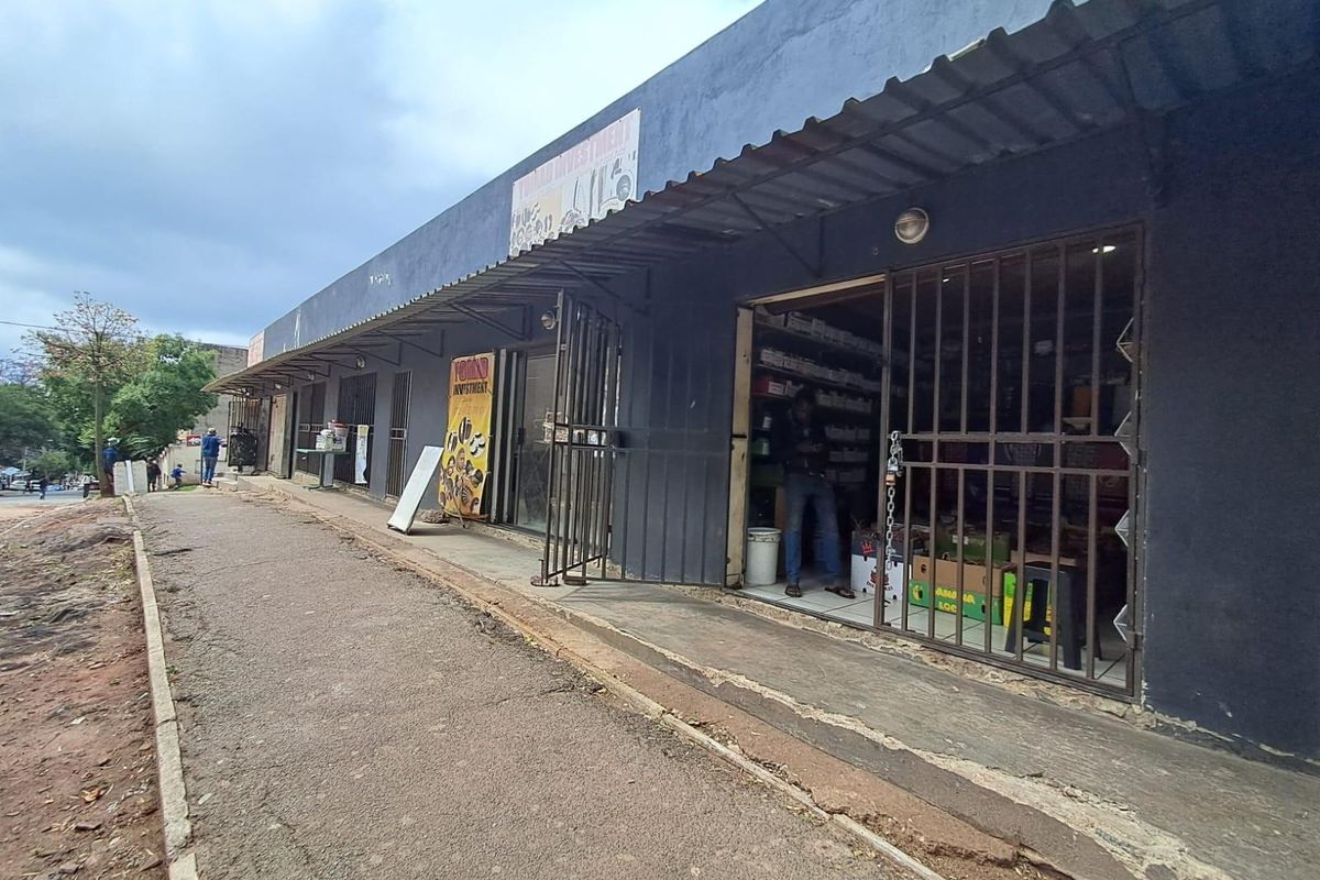 Front exterior view of a commercial strip building featuring multiple storefronts with metal security gates and a continuous corrugated metal awning.