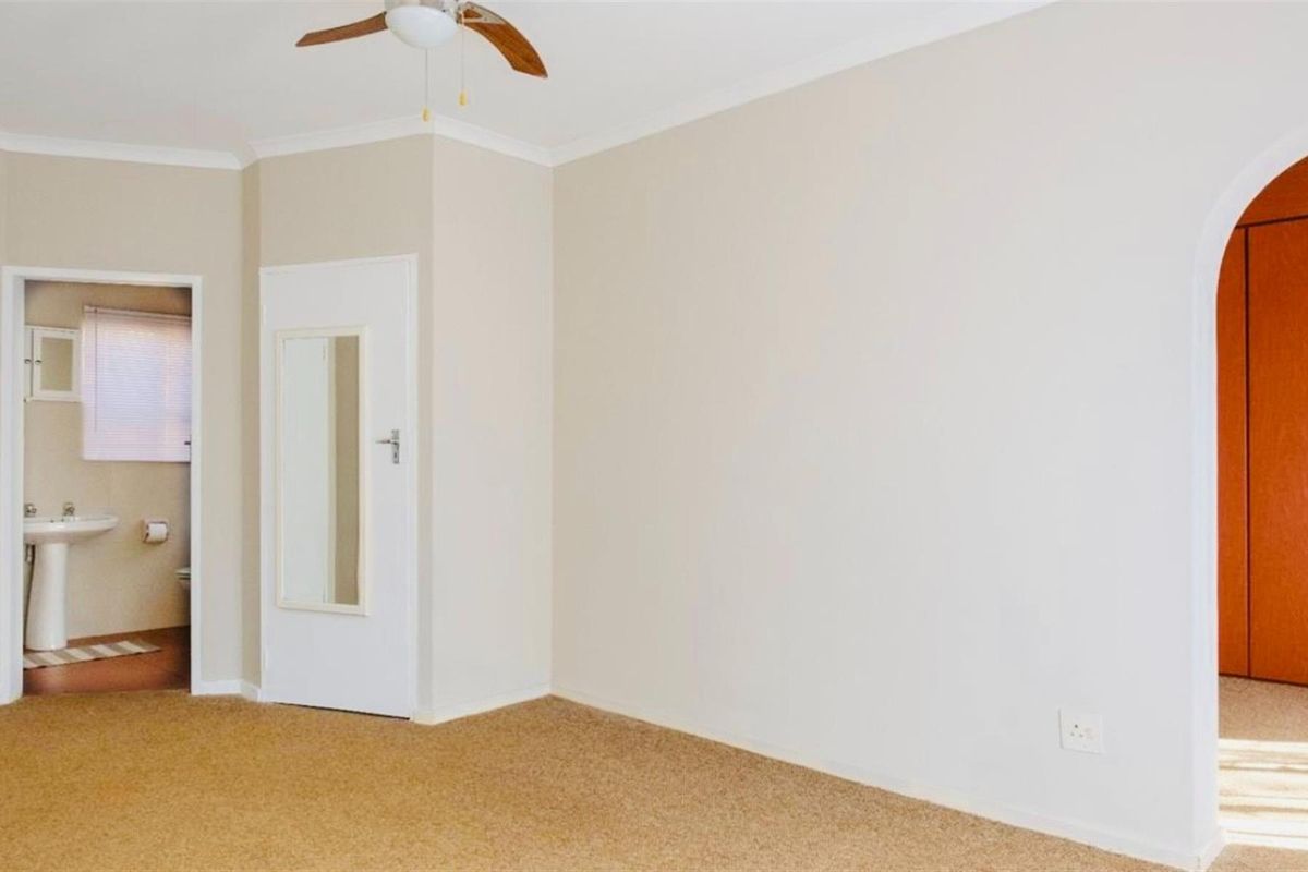 A spacious bedroom featuring neutral carpeting, a ceiling fan, and an arched doorway leading to an en-suite bathroom with a pedestal basin.