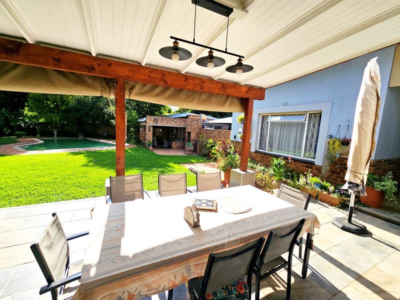 A covered outdoor patio area featuring paved flooring and timber beams, overlooking a private garden and swimming pool.