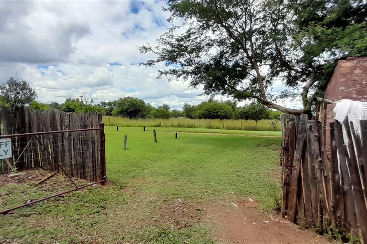 A rustic farm entrance featuring a wire gate, wooden pole fencing, and an adjacent brick outbuilding overlooking an open grassy field.