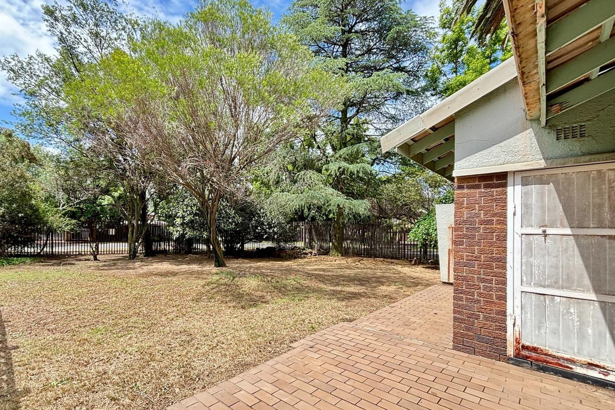 A rear garden area featuring a brick-paved patio, mature trees, and perimeter fencing, showing signs of weathering and maintenance needs.