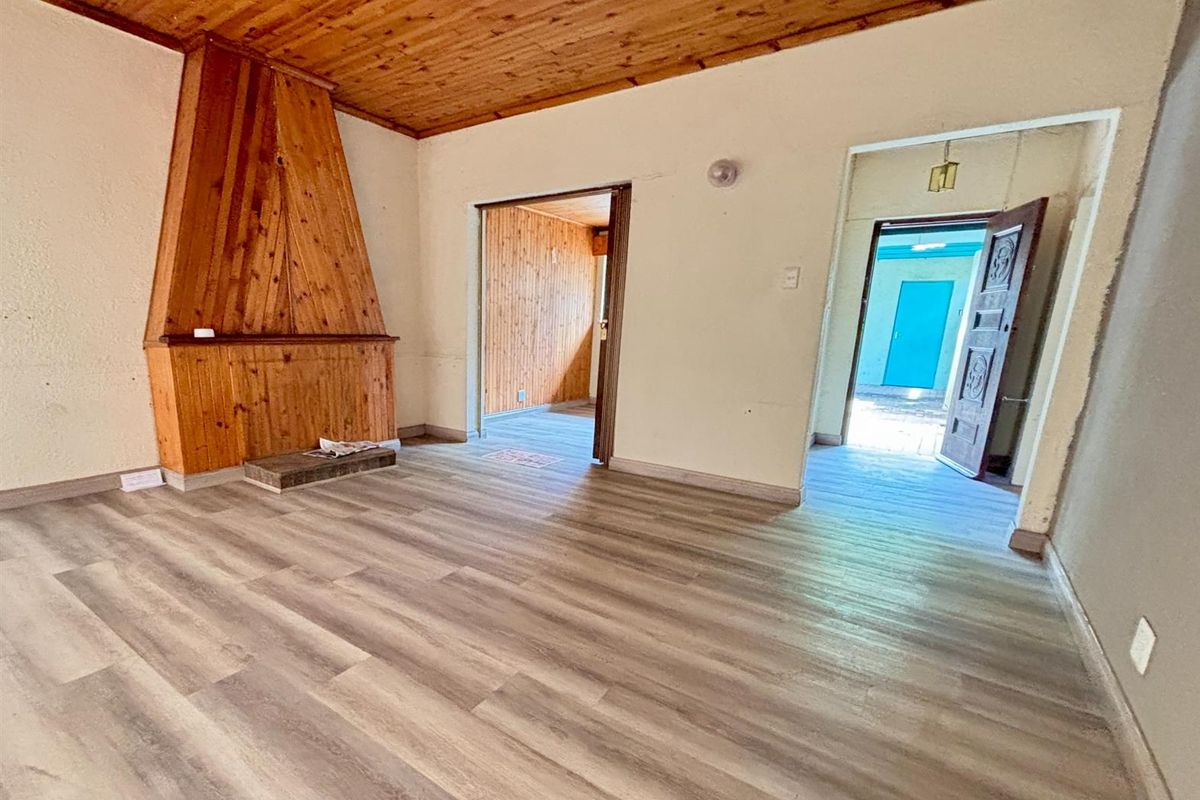 A living room featuring a prominent timber-clad fireplace, wood-paneled ceiling, and modern laminate flooring, with access to adjacent rooms and an exterior entrance.