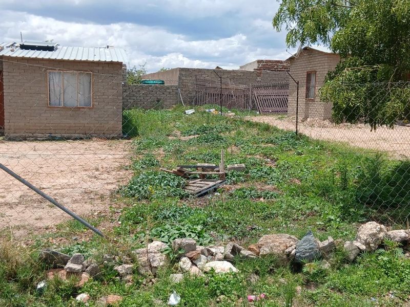 Exterior view of a residential property featuring multiple detached brick structures, a large unpaved yard, and perimeter chain-link fencing.