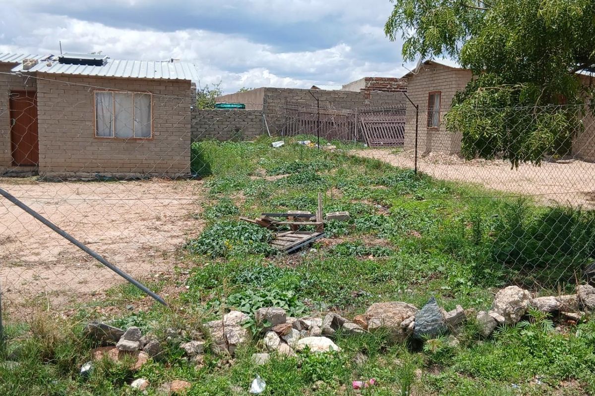 Exterior view of a residential property featuring multiple detached brick structures, a large unpaved yard, and perimeter chain-link fencing.