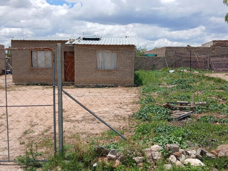 A modest single-story detached house featuring exposed brick construction, a corrugated metal roof, and a large, undeveloped yard enclosed by wire fencing.
