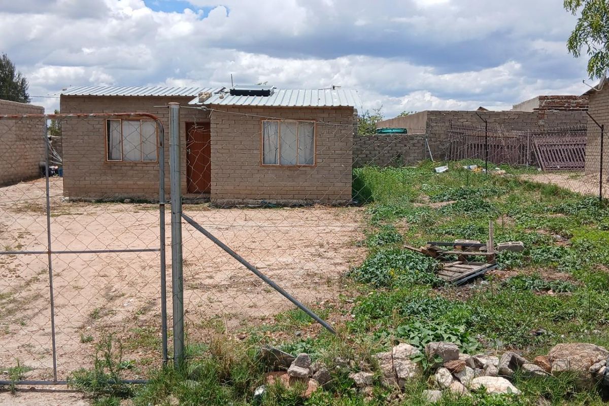 A modest single-story detached house featuring exposed brick construction, a corrugated metal roof, and a large, undeveloped yard enclosed by wire fencing.