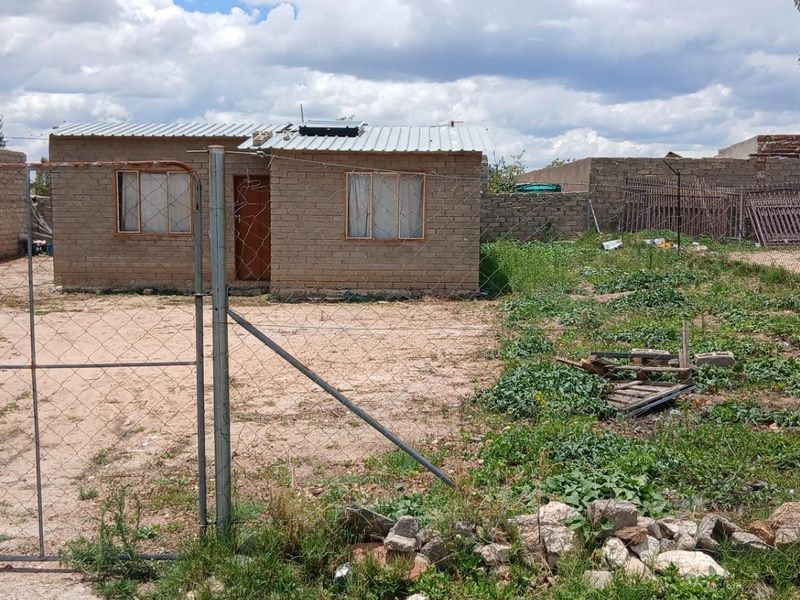 A detached single-story house featuring exposed brick masonry, a metal roof with a solar geyser, and a large, undeveloped plot with perimeter fencing.