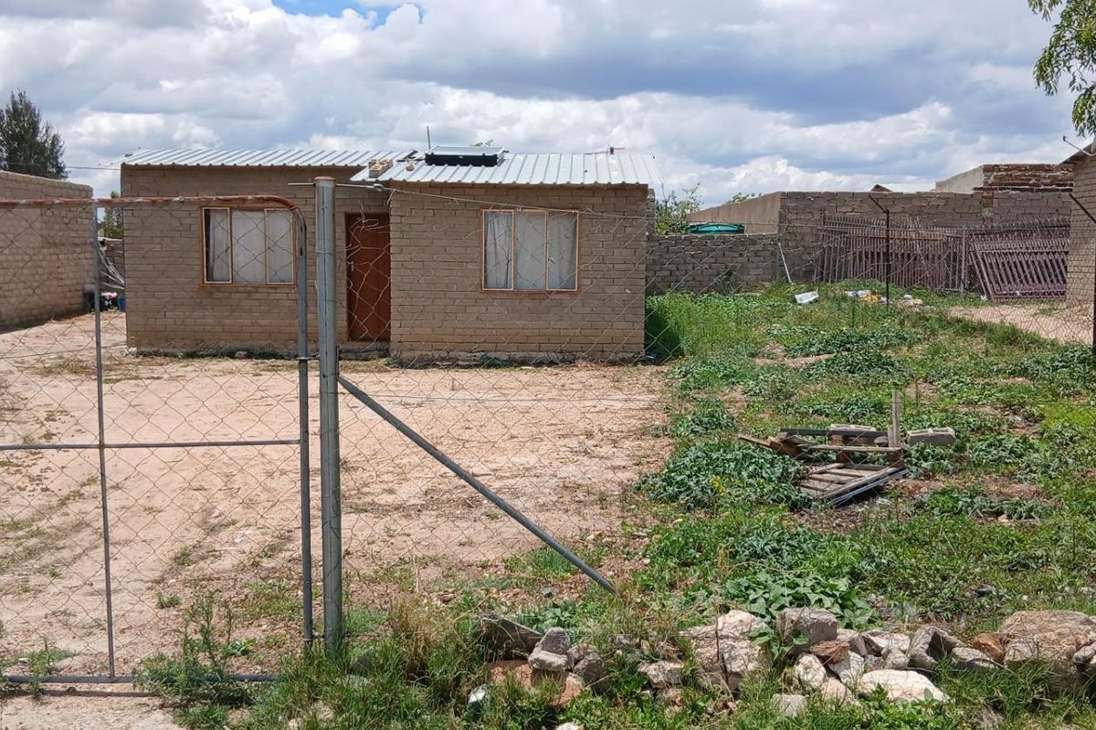 A detached single-story house featuring exposed brick masonry, a metal roof with a solar geyser, and a large, undeveloped plot with perimeter fencing.