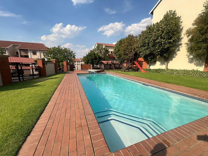 A well-maintained communal swimming pool area featuring brick paving, landscaped lawns, and perimeter walling within a residential apartment complex.