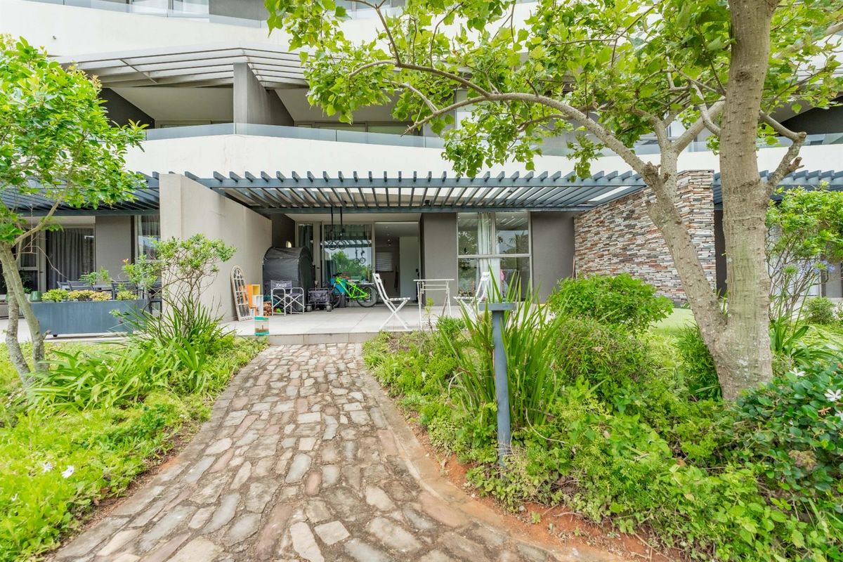 A ground floor apartment exterior featuring a paved walkway, landscaped garden, and a covered patio area with stone cladding and modern pergola.
