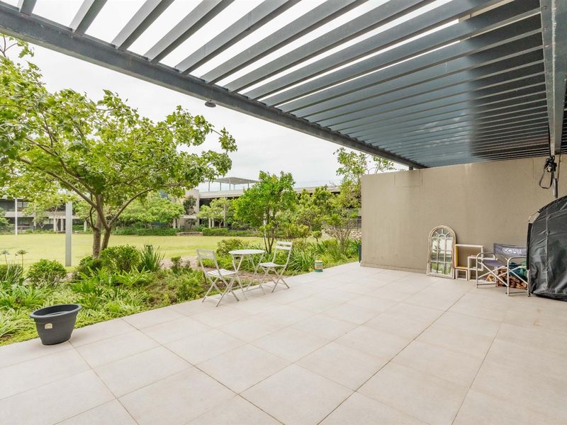 A spacious covered patio featuring tiled flooring and a modern metal pergola, overlooking a well-maintained communal garden area.