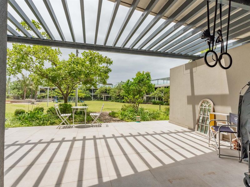 A spacious covered patio featuring a modern slatted pergola roof and tiled flooring, overlooking a landscaped communal garden area.